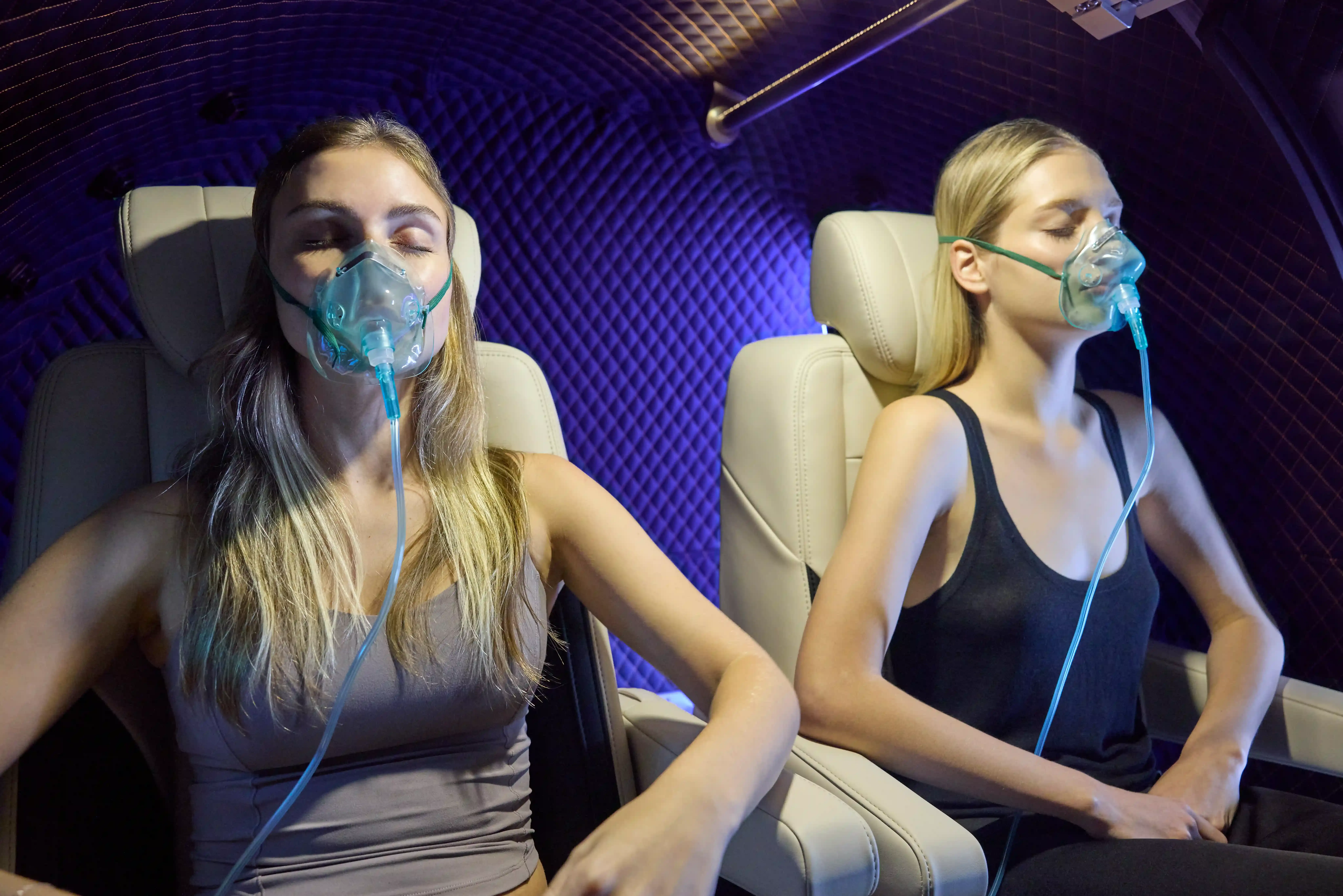 Two people seated inside a hyperbaric chamber, breathing oxygen through masks with their eyes closed