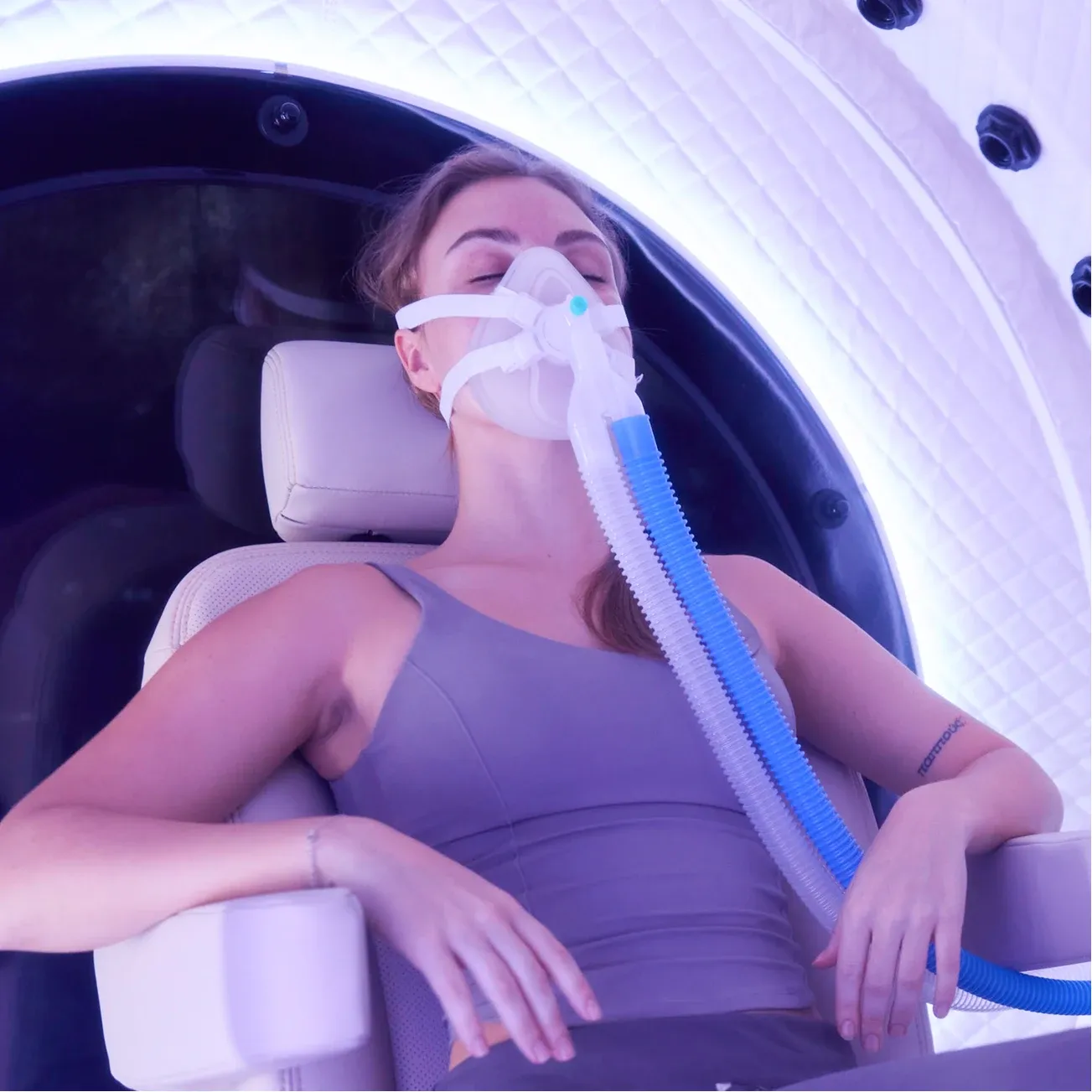 A woman reclined in the seat inside a Superhuman chamber, breathing through an oxygen mask