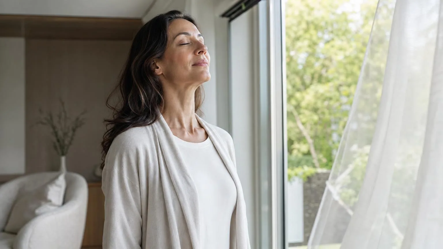 Woman breathing fresh morning air by an open window, sheer curtains moving softly, green foliage outside